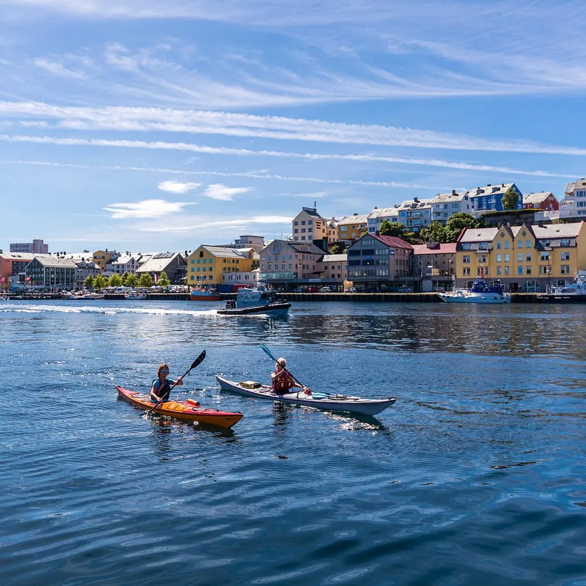 To personer padler kajakk i havnen foran fargerike bygninger og båter