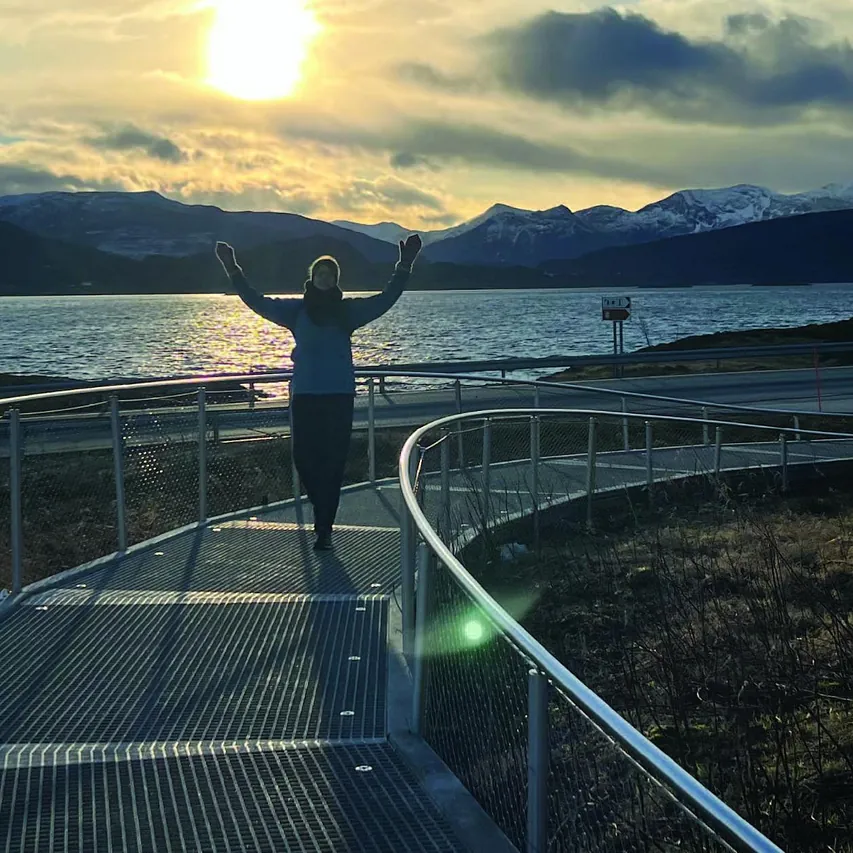 Person går langs oppbygd sti med armene opp. Solnedgang og fjell i bakgrunnen